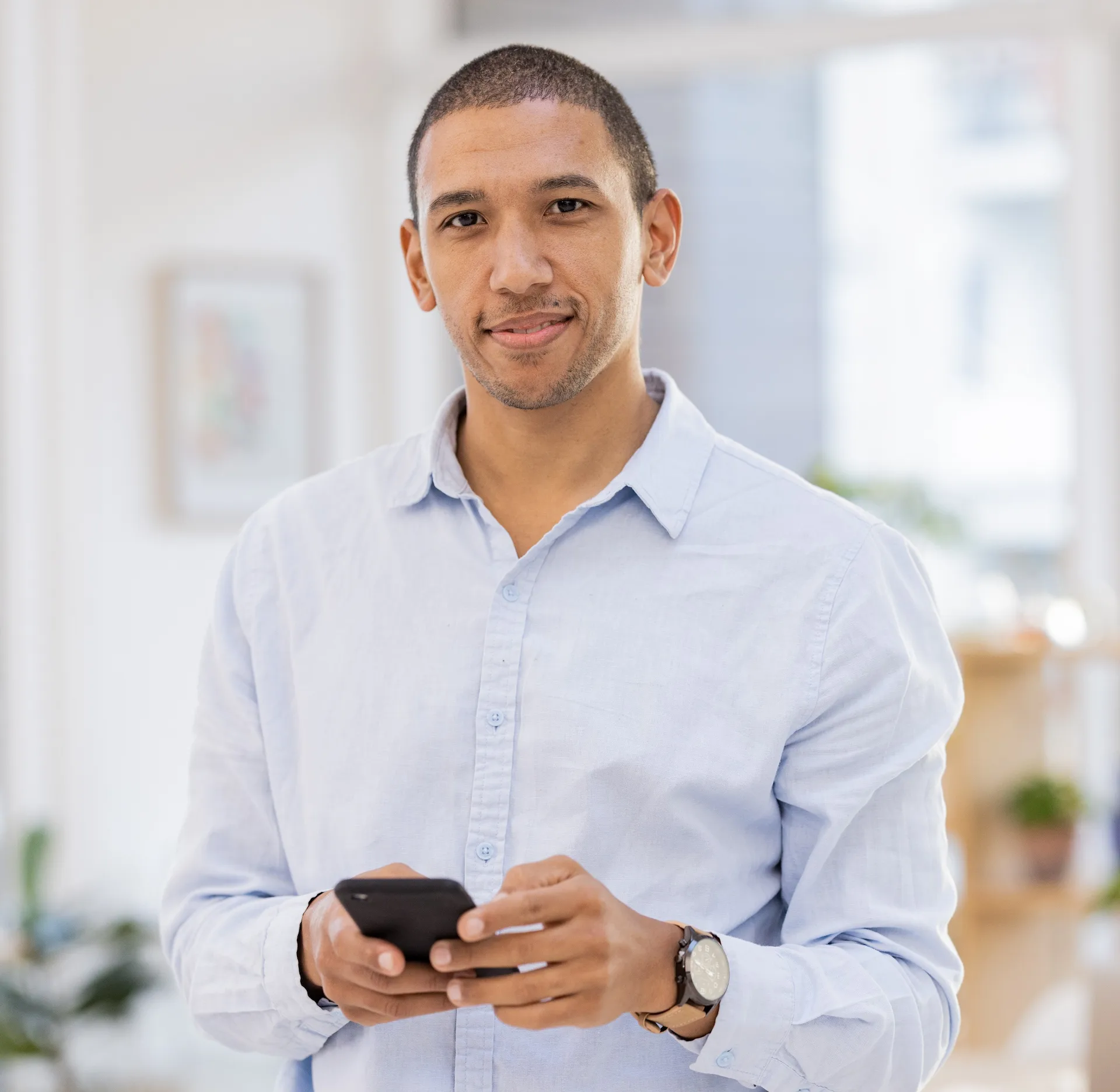 Man in light blue shirt holding a smartphone and smiling in a bright indoor setting.