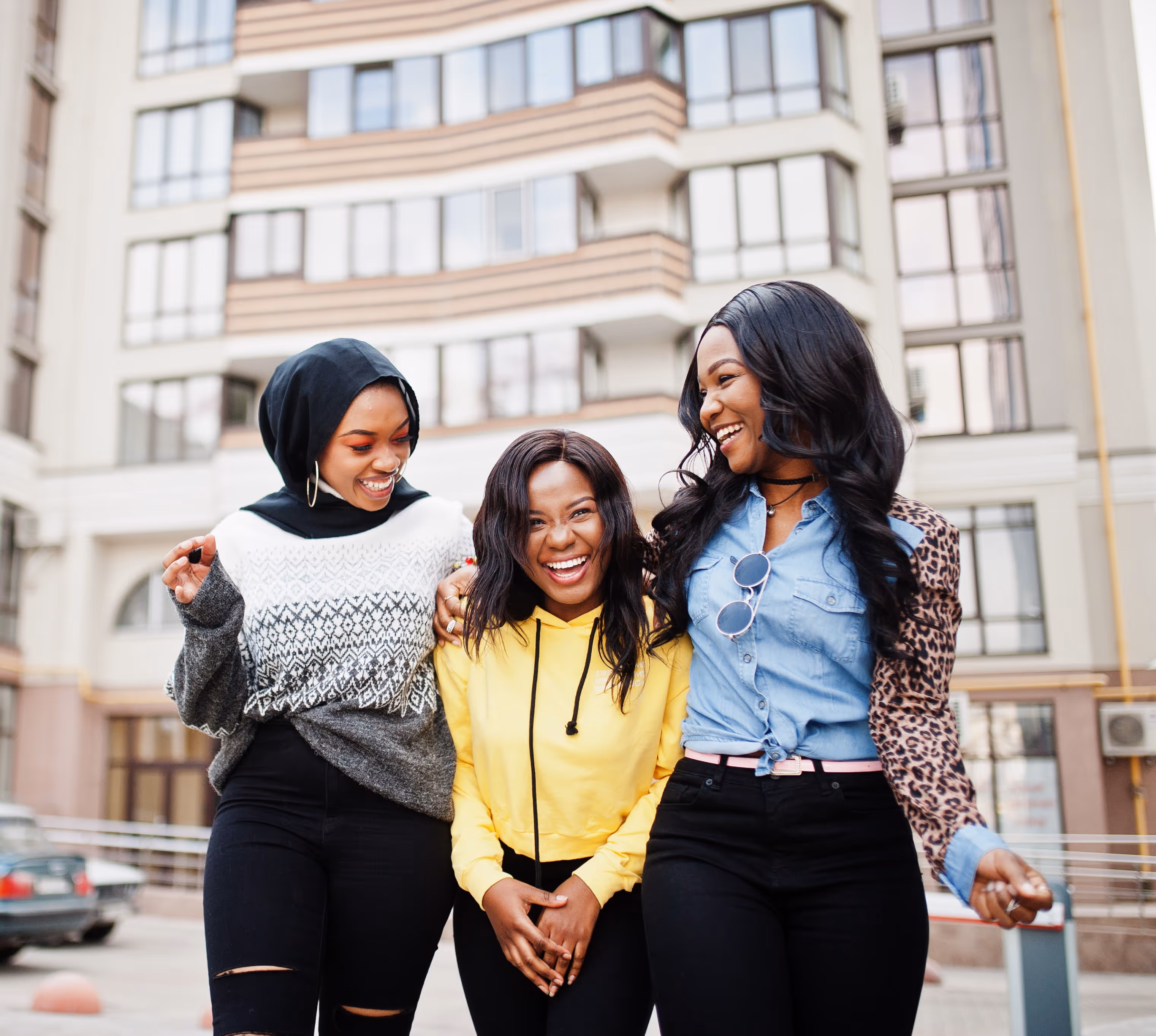 Three young women laughing and walking together in an urban setting in front of a modern building.
