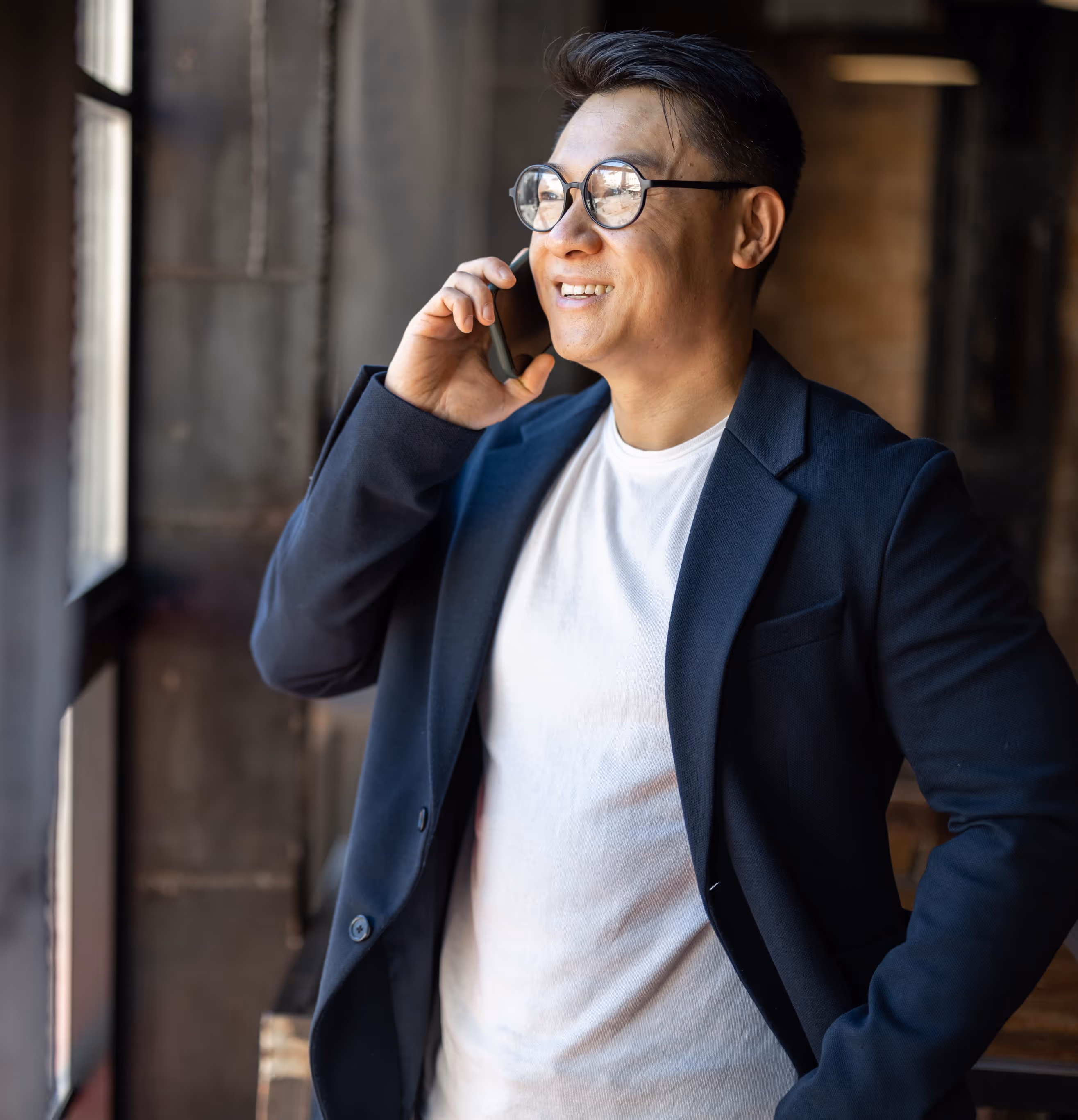 Smiling man wearing glasses and a dark blazer talking on a phone while looking out a window.