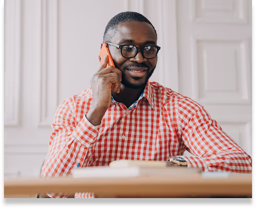 Smiling man wearing glasses and red checkered shirt talking on a phone while sitting at a desk.