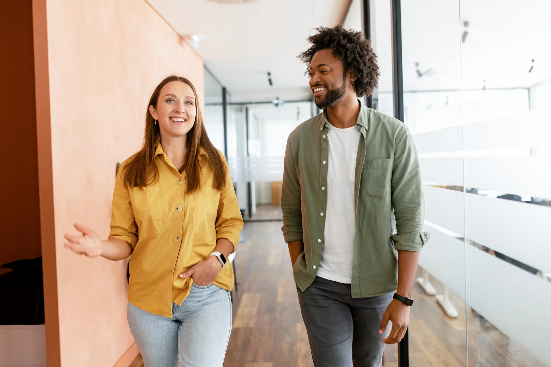 Two young colleagues, a woman in a yellow shirt and a man in a green shirt, walking and smiling in a modern office hallway.