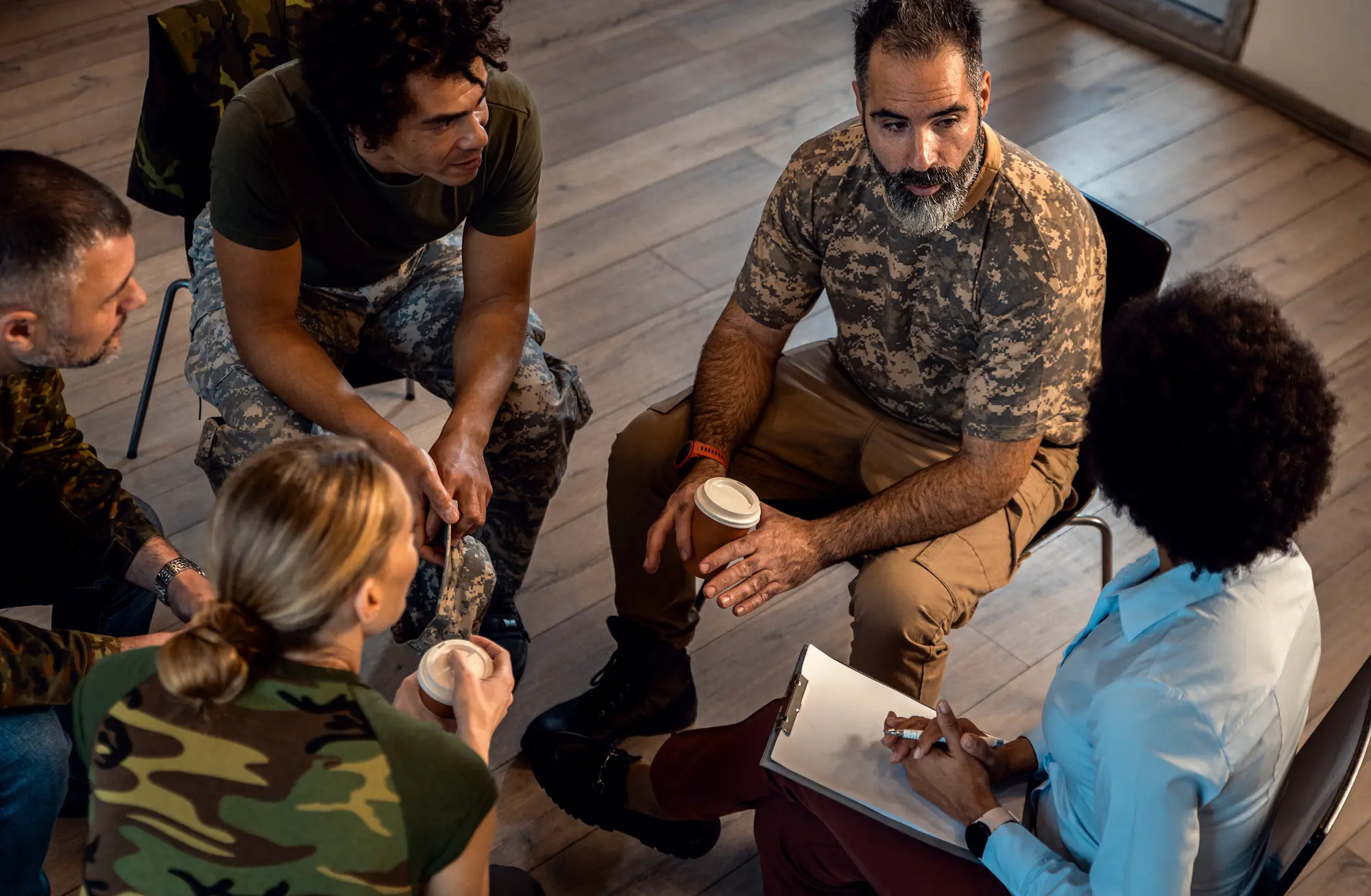 Group of military personnel in camouflage and a woman with a clipboard having a discussion seated in a circle.