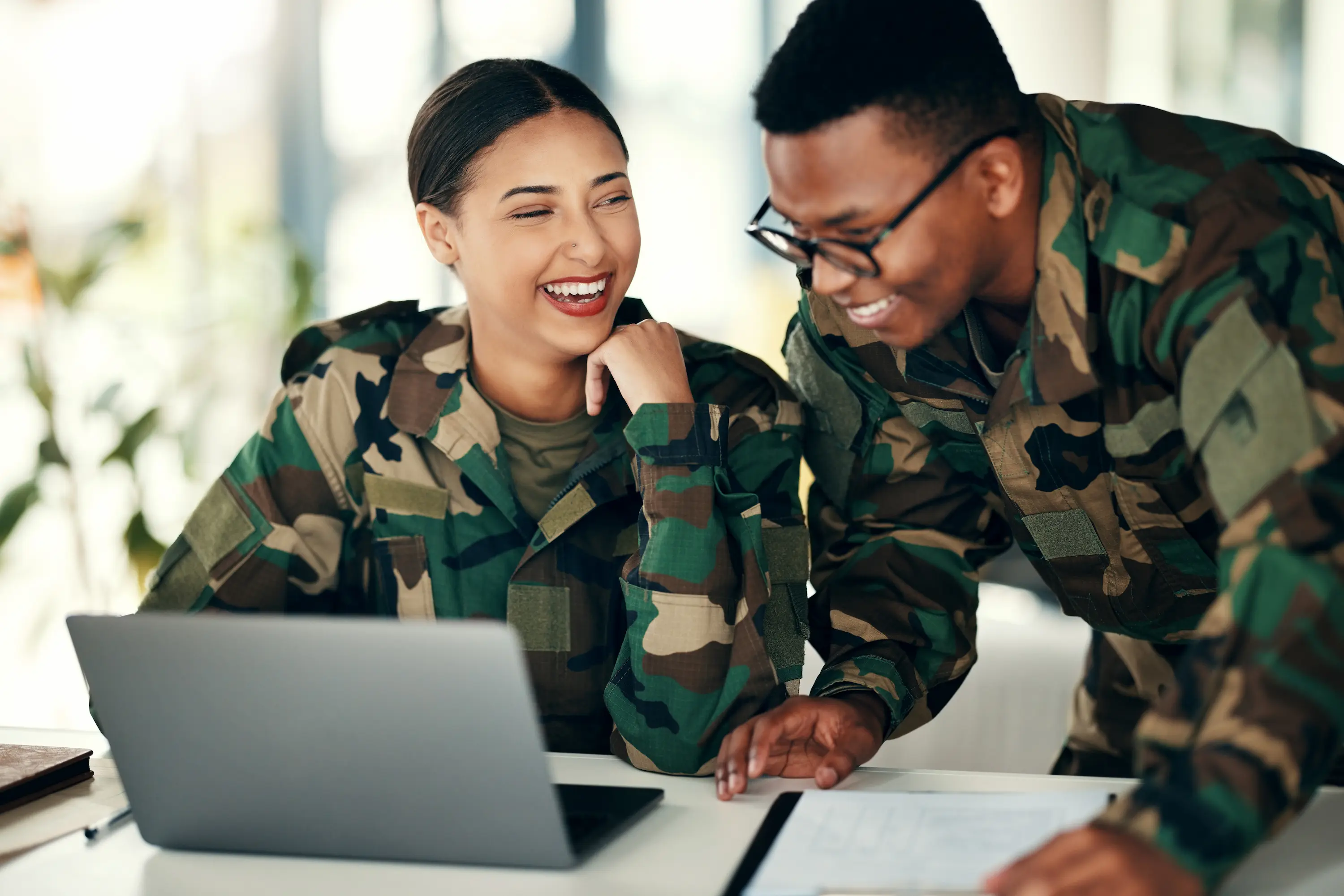 Two soldiers in camouflage uniforms smiling and working together at a laptop and documents.