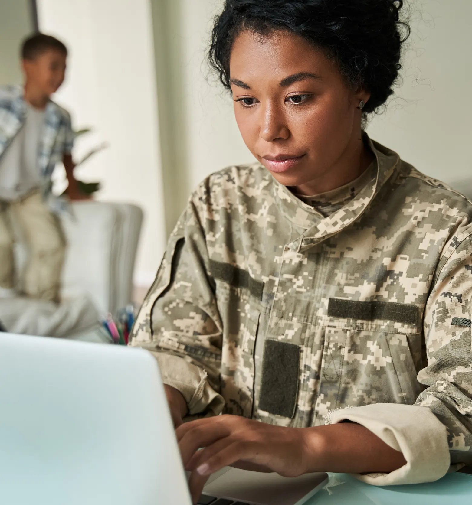 Woman in military uniform working on a laptop with a blurred child in the background.