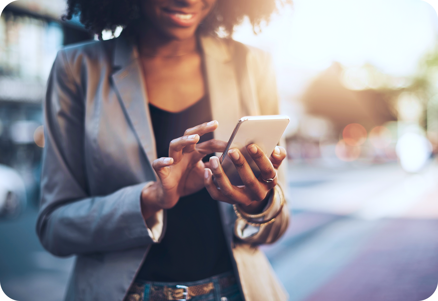 Woman in blazer smiling while using a smartphone outdoors in the sunlight.