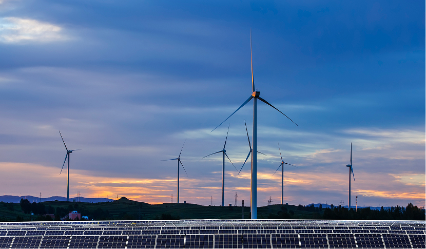Solar panels in front of multiple wind turbines under a cloudy sky at sunset.