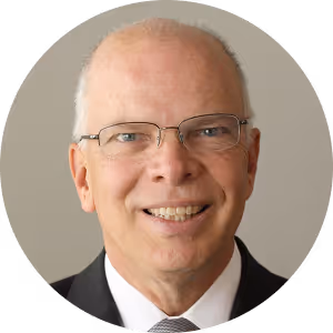 Smiling elderly man with glasses, wearing a black suit and gray tie, on a neutral background.