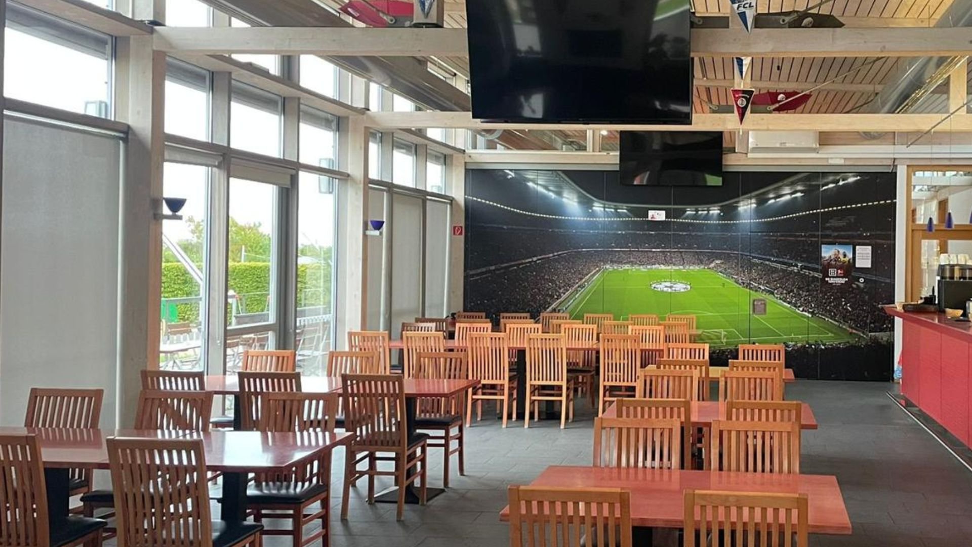Innenbereich des Restaurants „Zum Kelten“ mit Holztischen und -stühlen, großen Fenstern auf der linken Seite, Fernsehern unter der Decke und einem Wandbild, das ein Fußballstadion zeigt.
