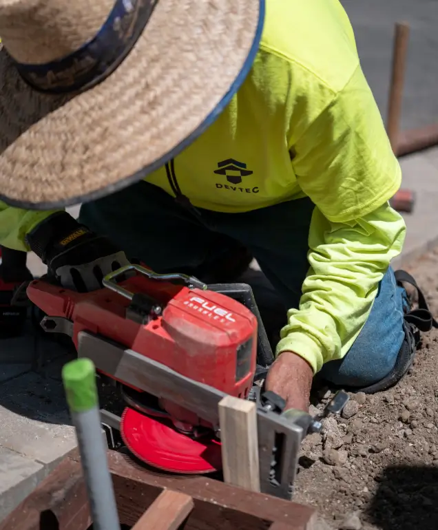 Construction worker in a straw hat and neon green shirt operating a red circular saw to cut wooden beams at a foundation site.