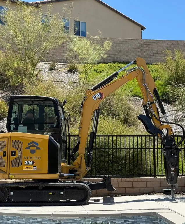 Yellow Caterpillar 304 mini excavator with hydraulic breaker attachment parked near a residential backyard pool.
