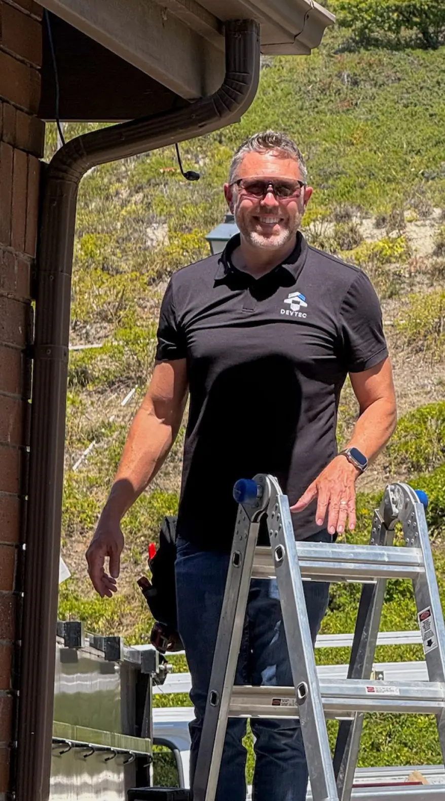 Smiling man wearing sunglasses and a black Devtec polo shirt standing next to a metal ladder outdoors.
