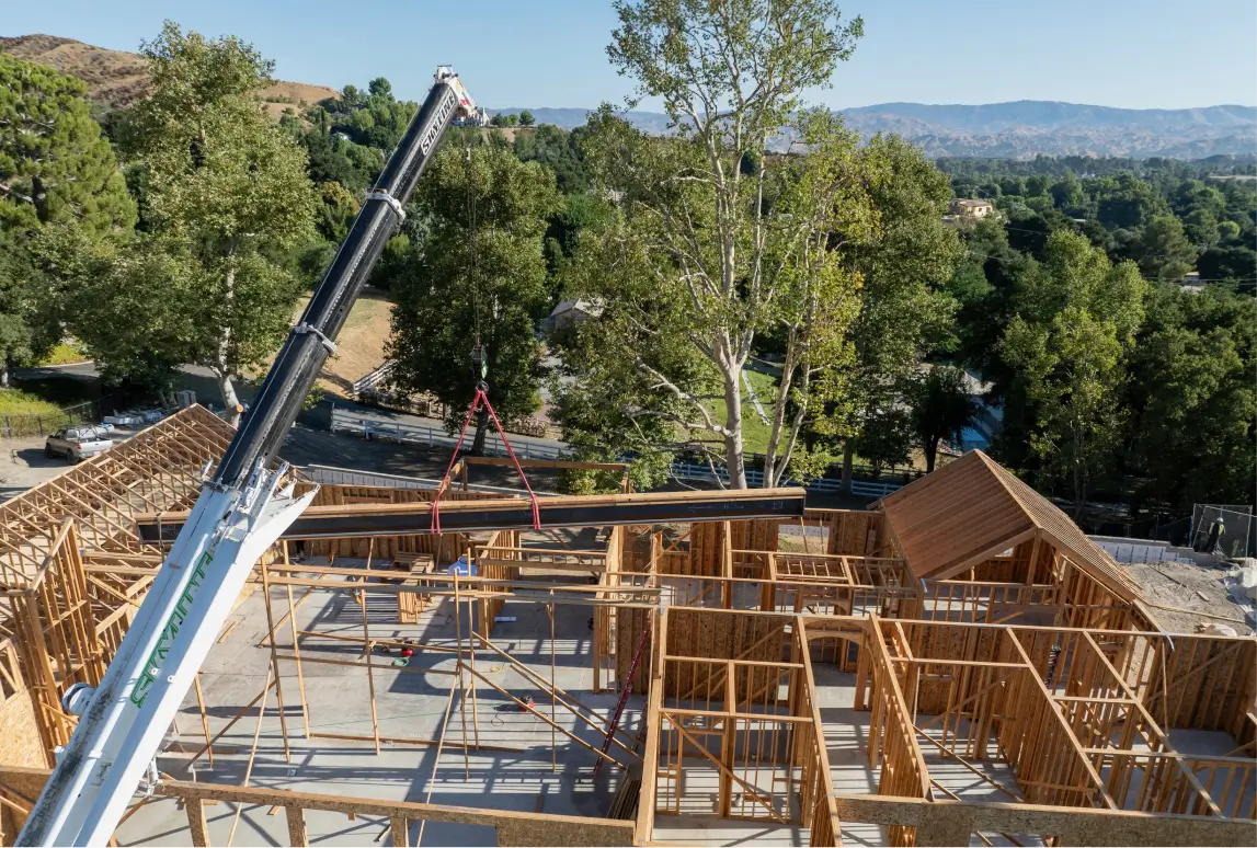 Crane lifting a large wooden beam over the wooden frame of a house under construction surrounded by trees and hills.