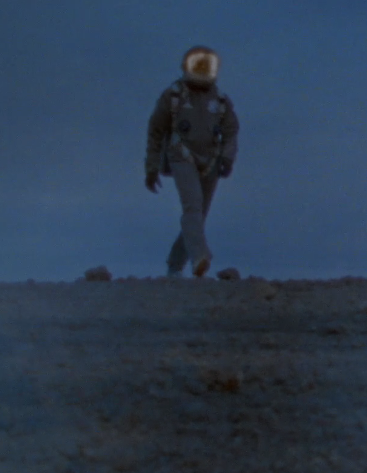 Astronaut in a space suit walking on a rocky surface under a dark, dusky sky.