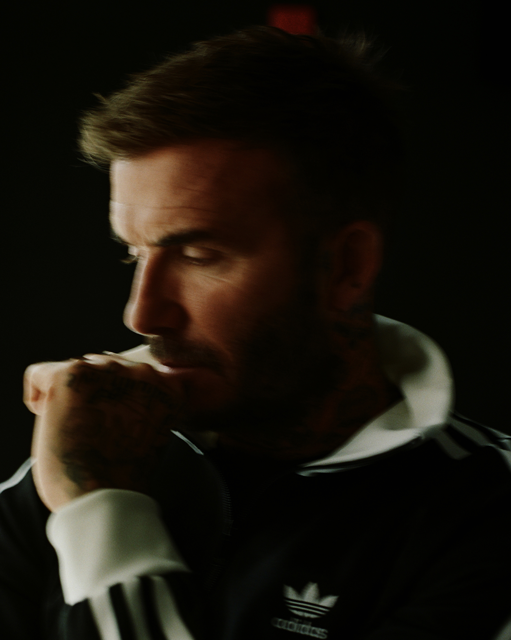 Man with tattoos wearing a black Adidas jacket with white stripes, resting his chin on his hand with eyes closed.