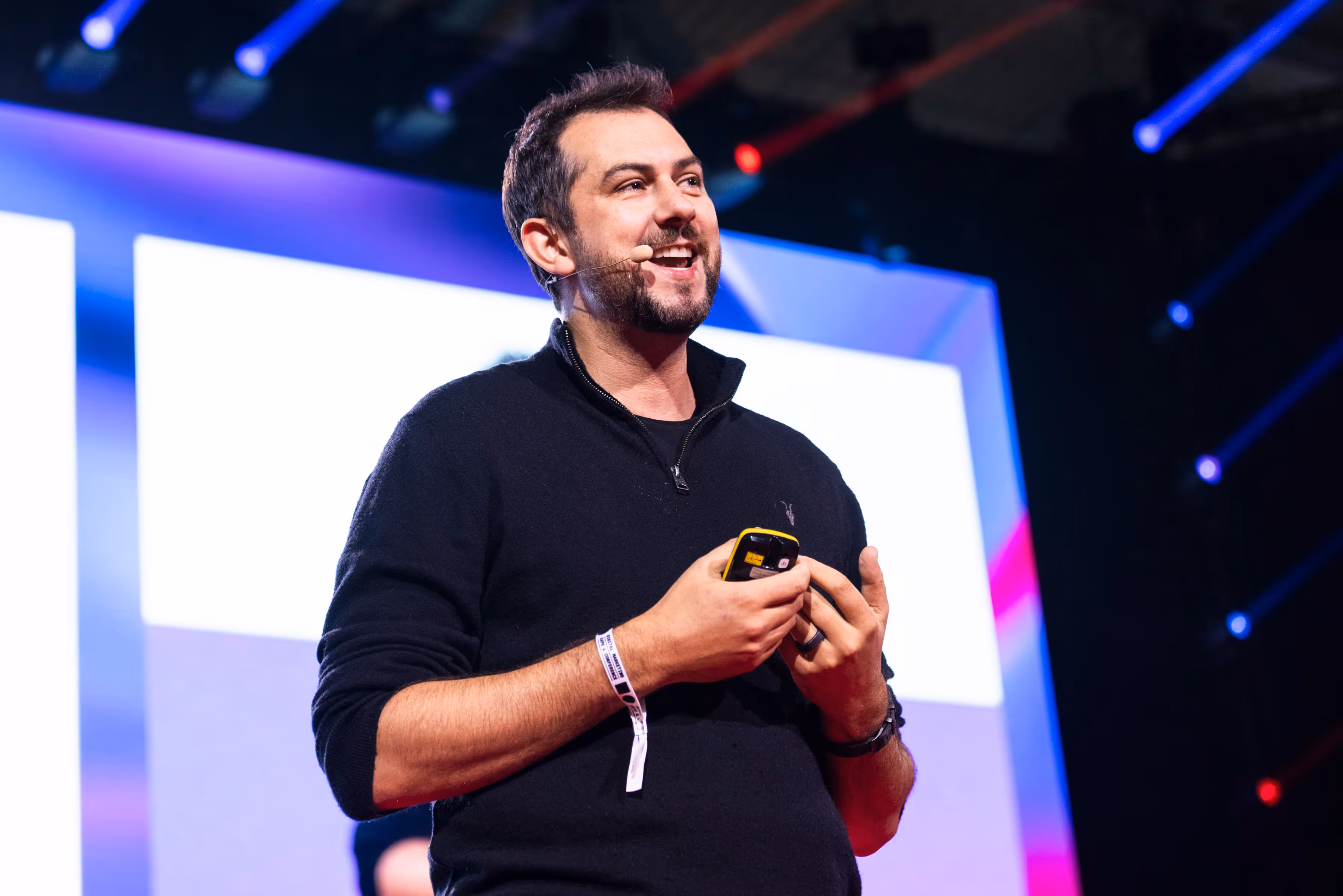 Daniel Hulme wearing a black sweater with a headset microphone speaking on stage, holding a yellow remote control.