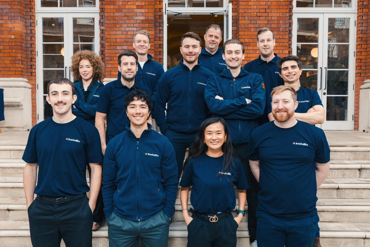 Group of thirteen people wearing matching navy Installio shirts and jackets posing on steps outside a brick building.