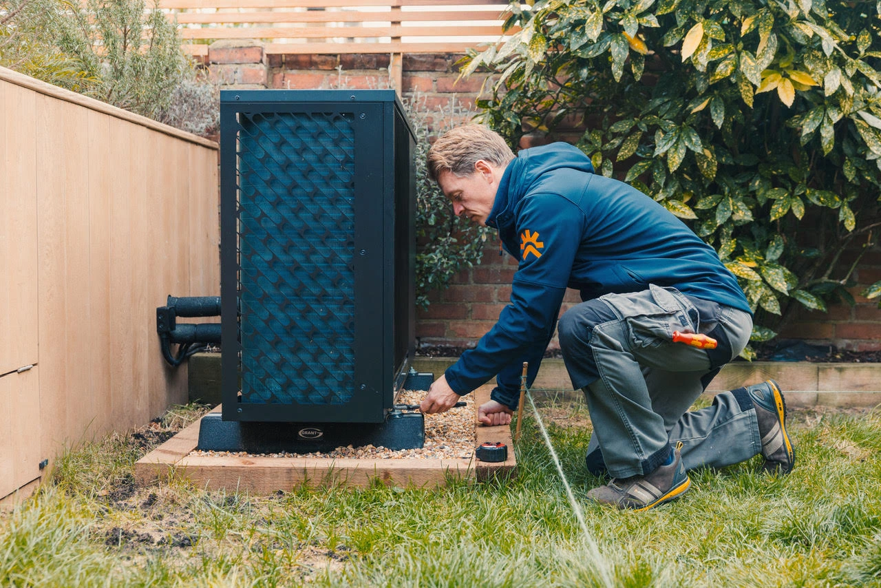 Technician in blue jacket installing or inspecting an outdoor heat pump unit in a backyard.