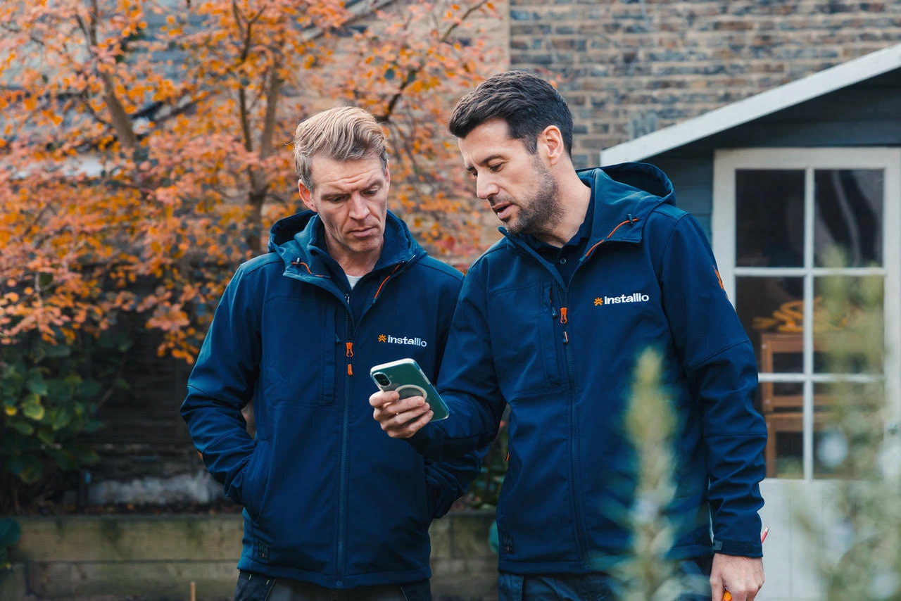 Two men wearing navy blue Installio jackets looking at a smartphone outdoors in a garden with autumn foliage.