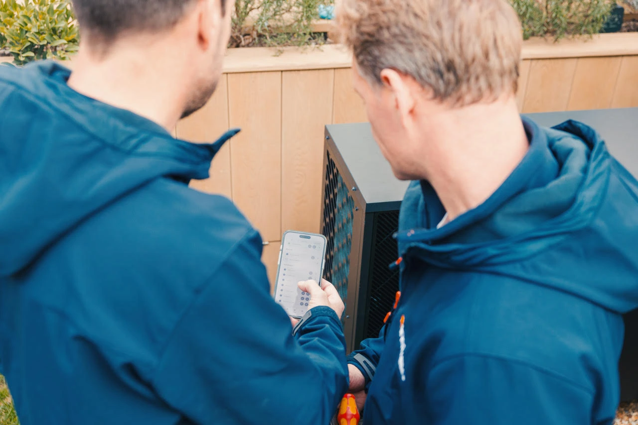 Two men in blue jackets looking at a smartphone screen while working on a piece of outdoor equipment.