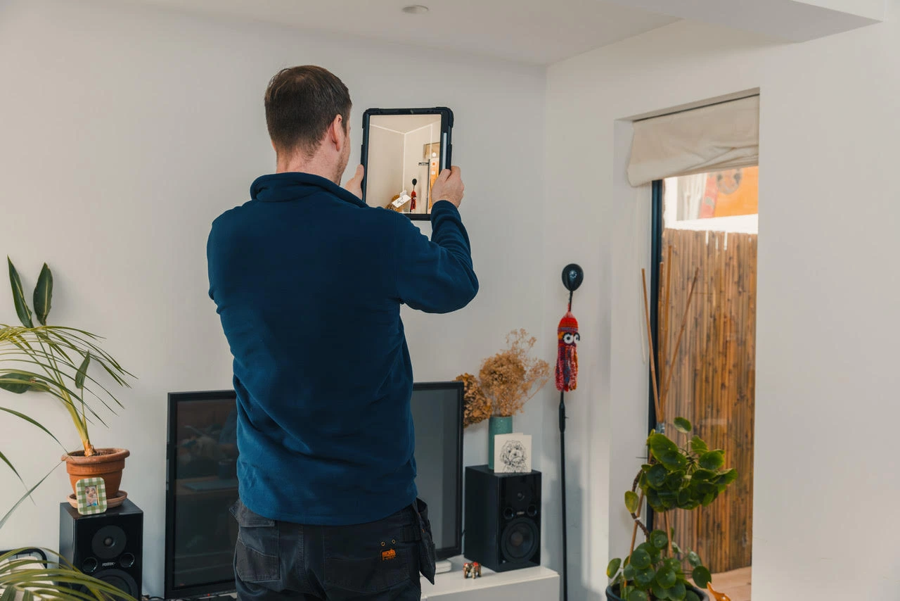 Man in blue jacket using a tablet to scan or measure a corner of a room with plants and speakers nearby.