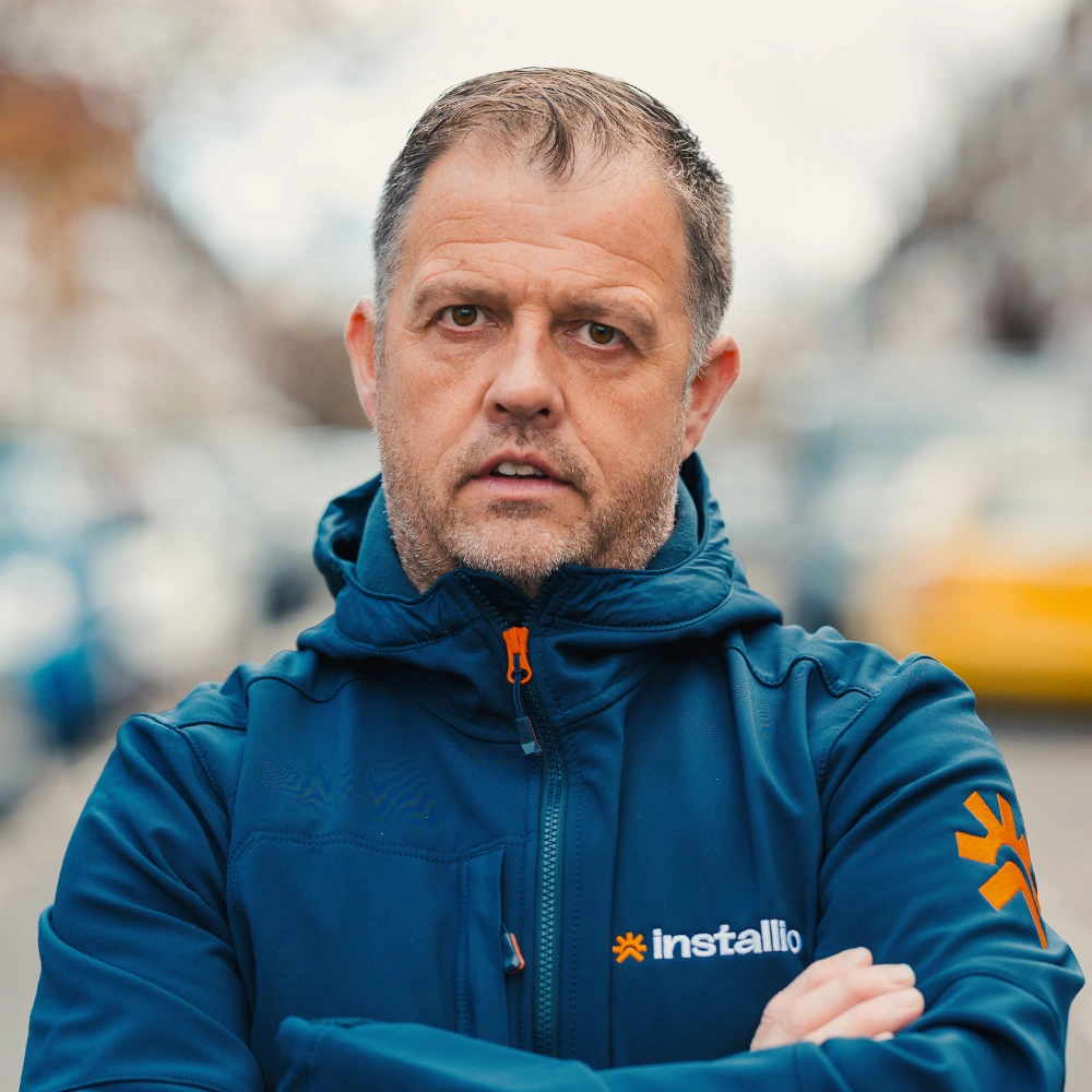 Man with short hair and stubble wearing a blue Installio jacket standing outdoors with arms crossed.