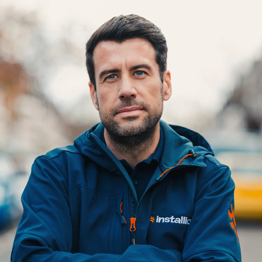 Man with dark hair and beard wearing a navy blue jacket with 'installio' logo, looking confidently at the camera outdoors.