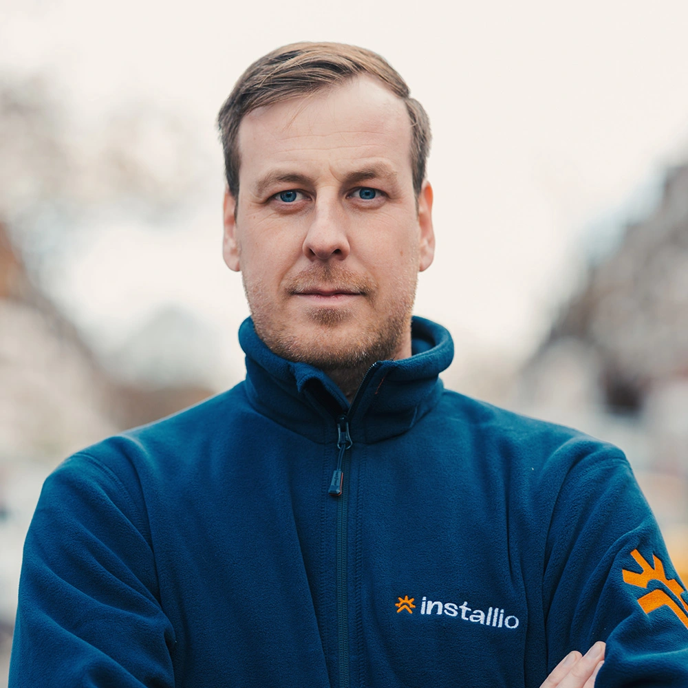 Man with short hair and blue eyes wearing a navy blue Installio fleece jacket, looking directly at the camera with arms crossed.