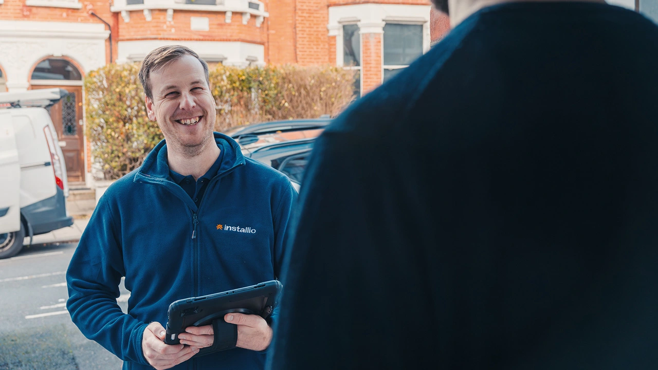 Smiling man in blue jacket holding a tablet, talking to another person outdoors in a residential street.