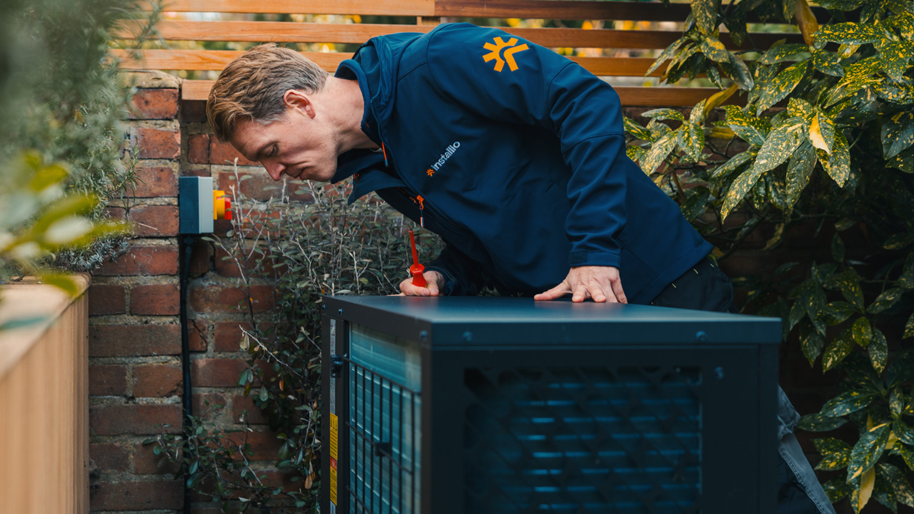 Technician in a blue jacket inspecting an outdoor HVAC unit near a brick wall and plants.