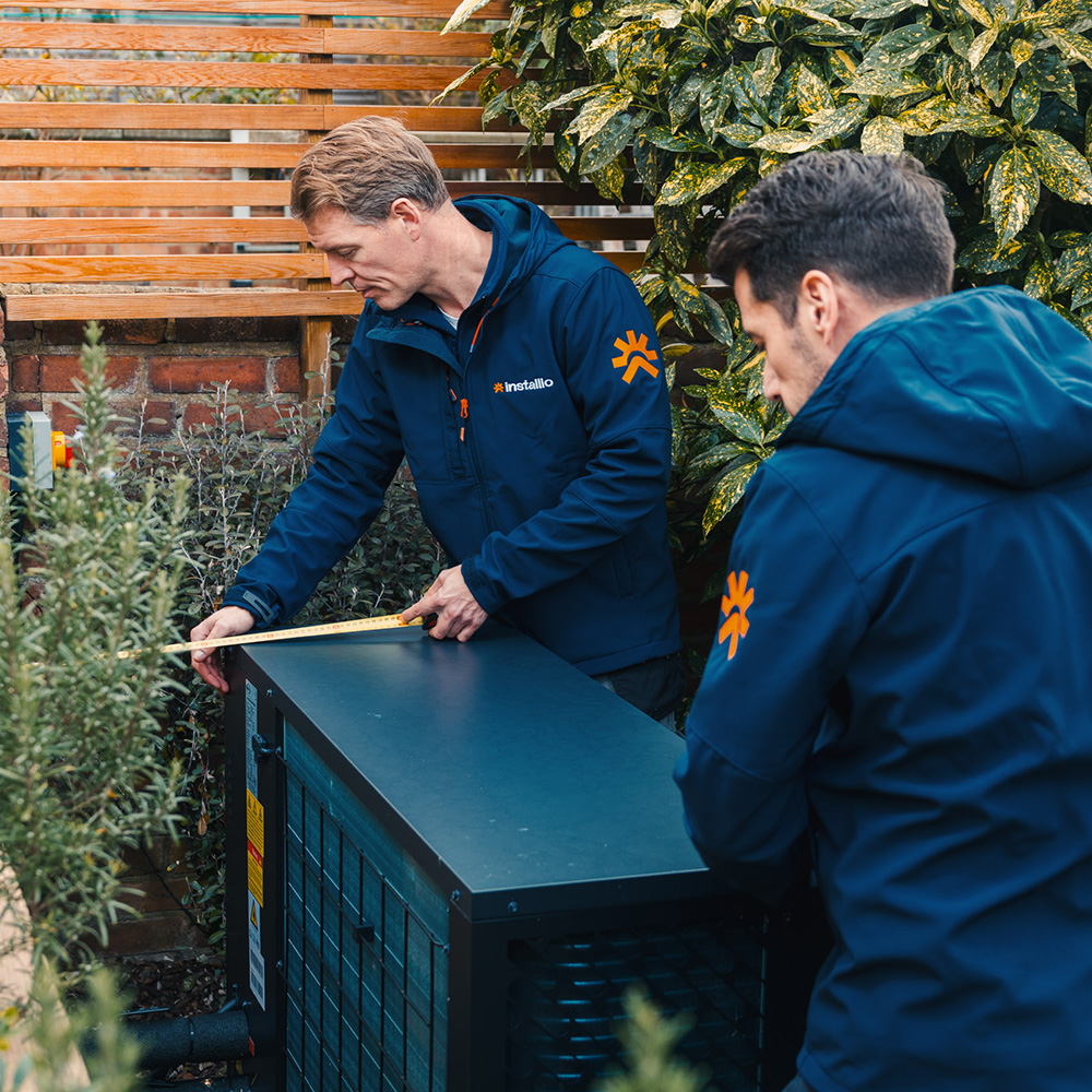 Two men in navy jackets measuring a large outdoor heat pump unit surrounded by plants and wooden slats.