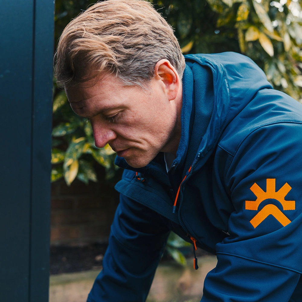 Man with short light brown hair wearing a blue jacket with an orange logo on the sleeve, looking down outdoors near greenery.