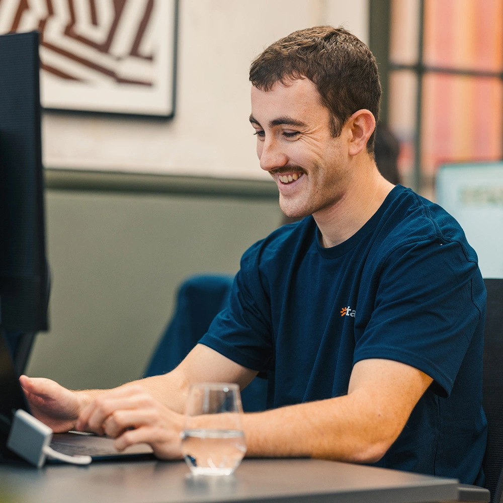Smiling man with a mustache working on a laptop at a desk with a glass of water.