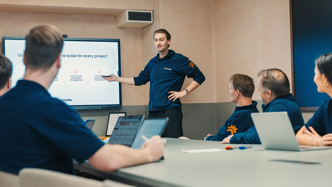 Man in navy jacket giving a presentation to four colleagues in a meeting room with laptops and a screen displaying project management points.