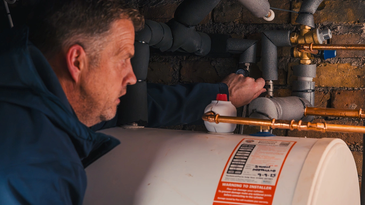 Man inspecting and adjusting pipes connected to a water heater in a basement with exposed brick walls.