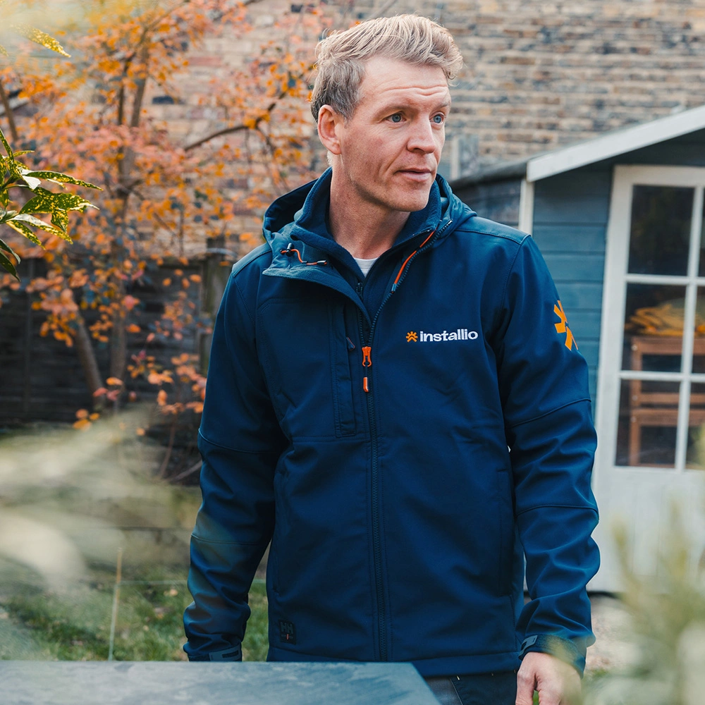 Man wearing a navy blue jacket with Installio logo stands outdoors near autumn foliage and a wooden shed.