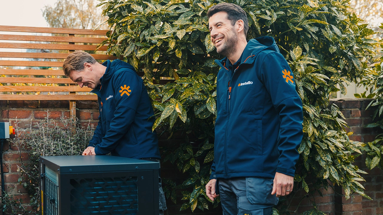 Two men wearing navy jackets with Installio logos working and smiling near an outdoor machine by a brick wall and green shrub.