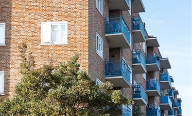 Brick apartment building with blue balconies and leafy tree in front under a clear sky.