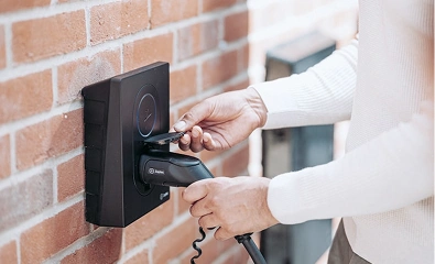 Person plugging an electric vehicle charger into a wall-mounted charging station on a brick wall.