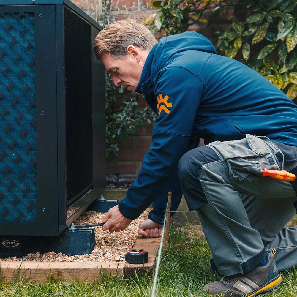 Technician in a blue jacket kneeling outdoors, using a wrench to adjust a black HVAC unit mounted on gravel and wood.