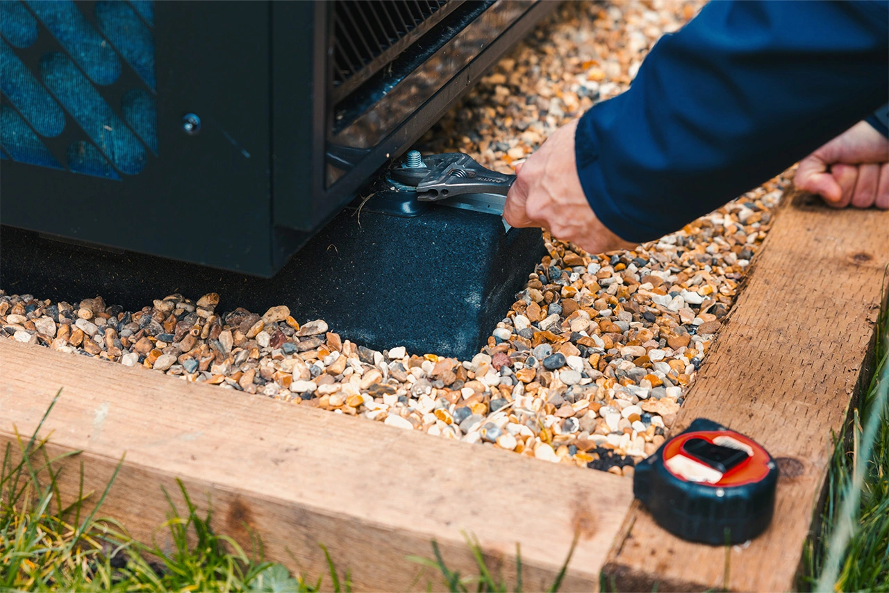 Person using a wrench to tighten a bolt on an outdoor unit placed on gravel and wooden frame.