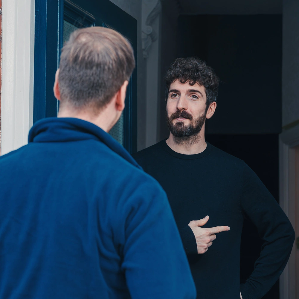 Bearded man in black shirt at a doorway pointing at a man in blue jacket facing him.