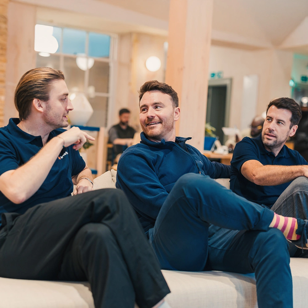 Three men casually talking and sitting on a couch in a modern office lounge.