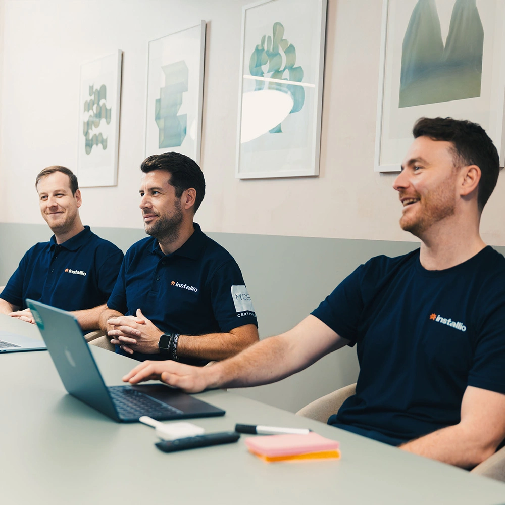 Three men wearing navy Installio shirts seated at a table with laptops, smiling and engaged in discussion in a meeting room.