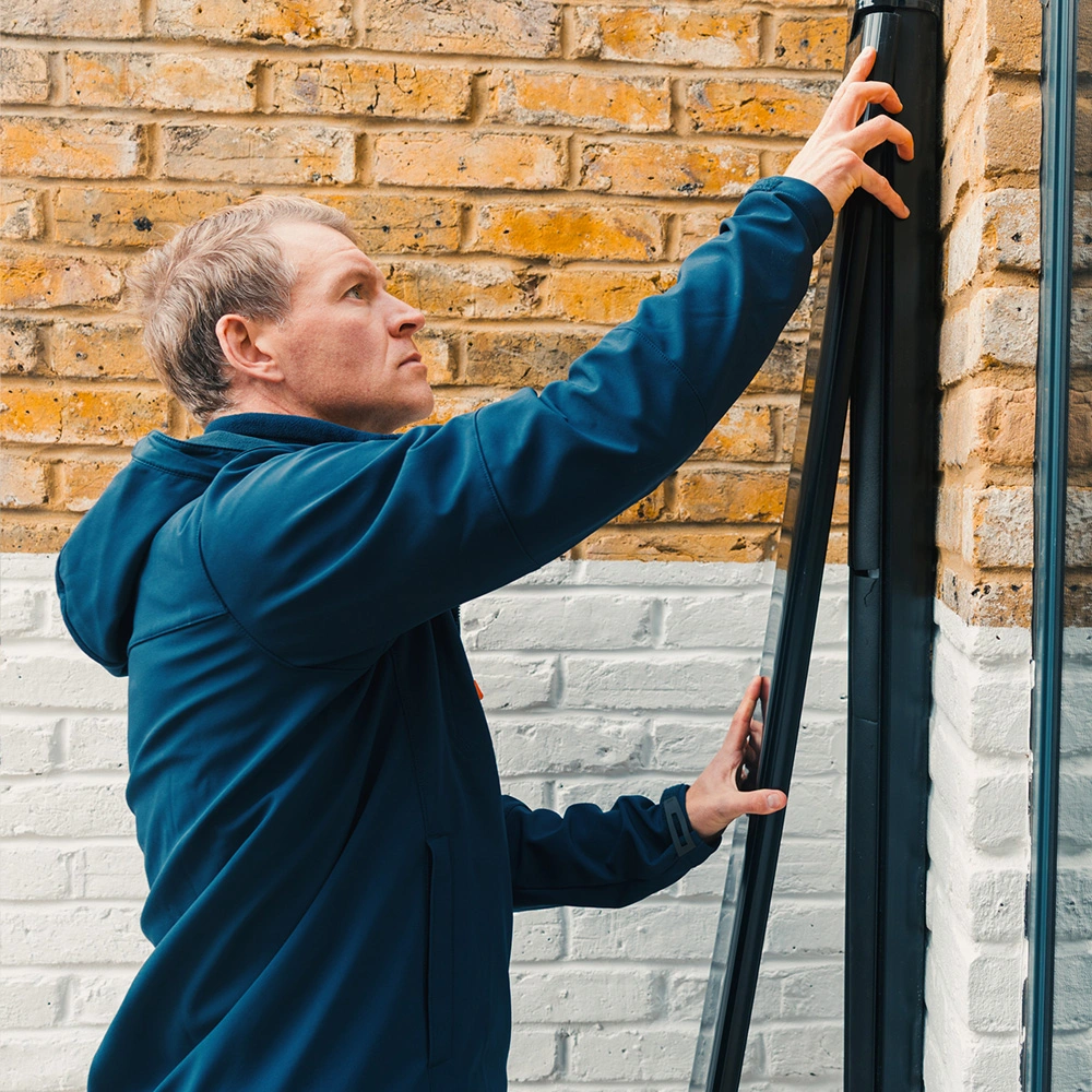 Man in a blue jacket installing or adjusting a black window frame against a brick wall.