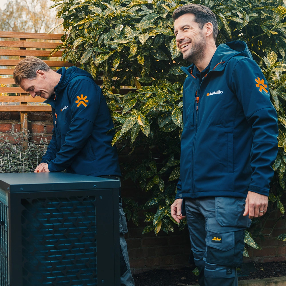 Two male technicians in blue Installio jackets working outdoors by a large air conditioning unit with greenery in the background.