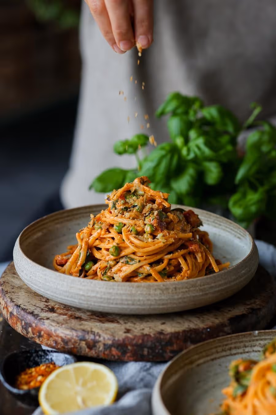 Hand sprinkling seasoning over a plate of spaghetti with peas and tomato sauce, garnished with herbs.