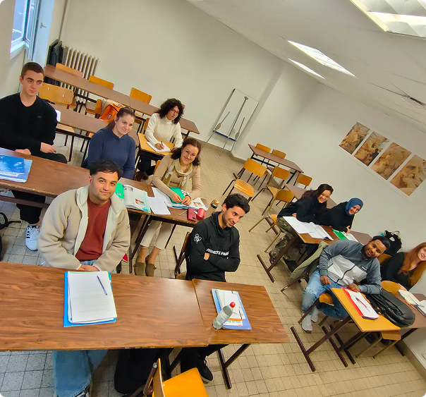 Groupe d'étudiants assis dans une salle de classe avec des bureaux en bois, prenant des notes et souriant vers l'avant.