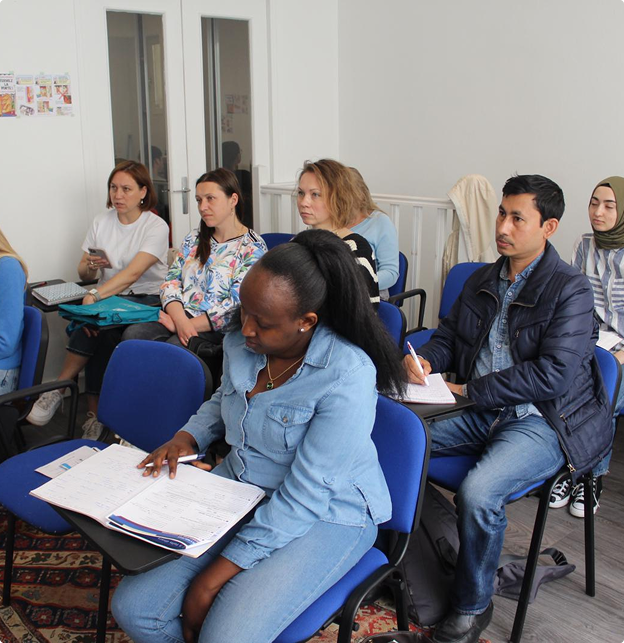 Un groupe diversifié d'adultes assis sur des chaises bleues dans une salle de classe, prenant des notes et écoutant attentivement.
