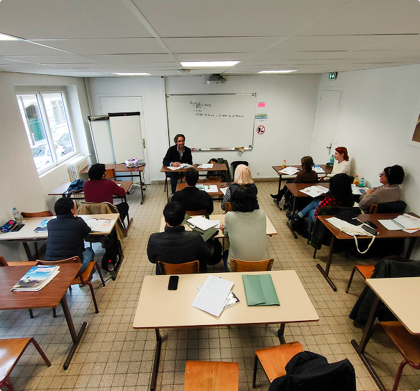 Salle de classe avec un enseignant parlant devant un tableau blanc et des étudiants assis à des bureaux.
