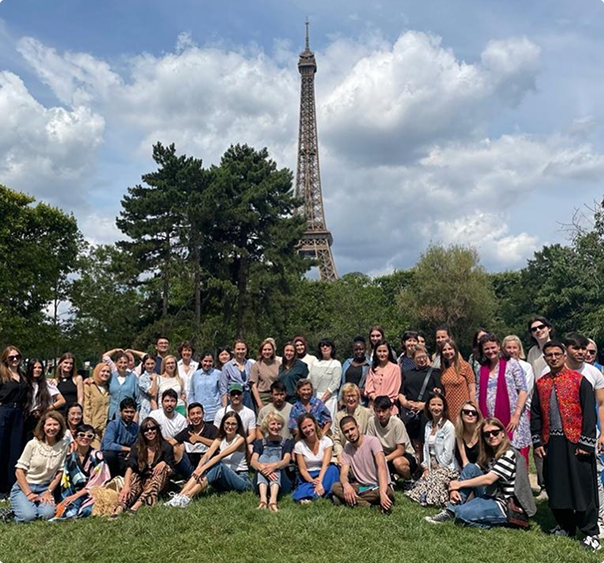 Groupe de personnes assises et debout sur l'herbe avec la tour Eiffel et des arbres en arrière-plan sous un ciel nuageux.